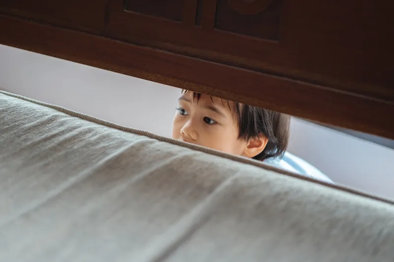 A young boy peeking out from behind a bed