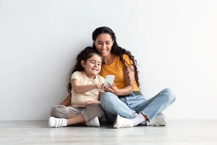 Online Shopping. Cute Little Girl And Her Mom Using Smartphone At Home, Happy Middle Eastern Mother And Preteen Daughter Relaxing On Floor With Mobile Phone, Enjoying Purchasing In Internet