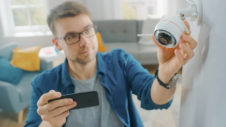 A man adjusting a Wi-Fi security camera on his apartment wall while checking the feed on his phone