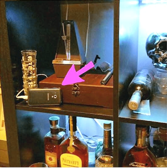 An old phone with a pink arrow pointing at it, charging on a shelf beside a wooden box and bottles.