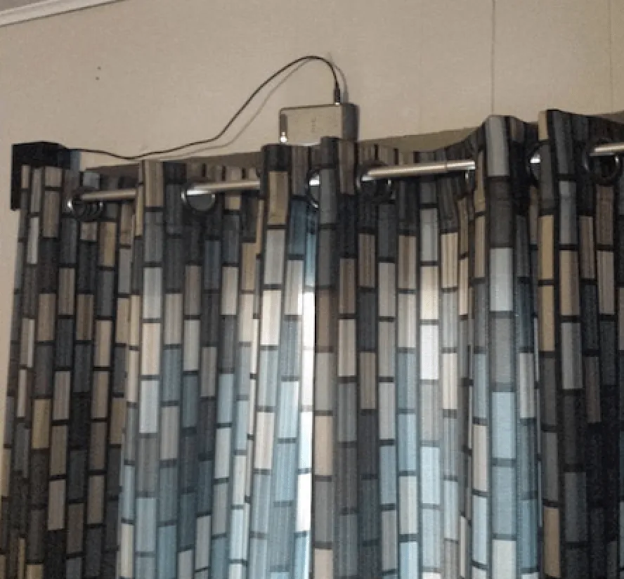 An old phone resting on a curtain rail next to a portable charger, used as a security camera