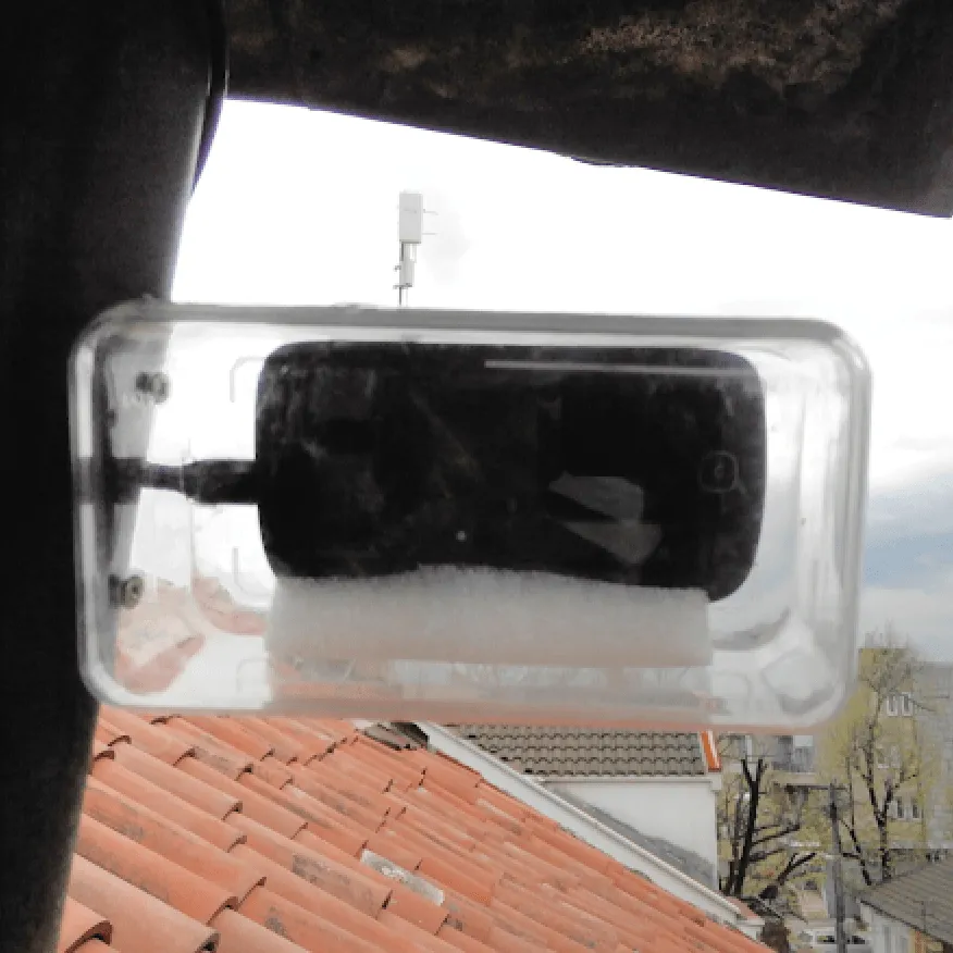 A phone with a charging cable sealed in a clear plastic box mounted at a window.