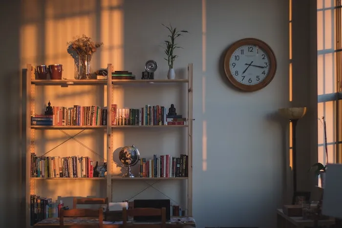 A wooden bookshelf full of books next to a wall clock in evening sunlight.