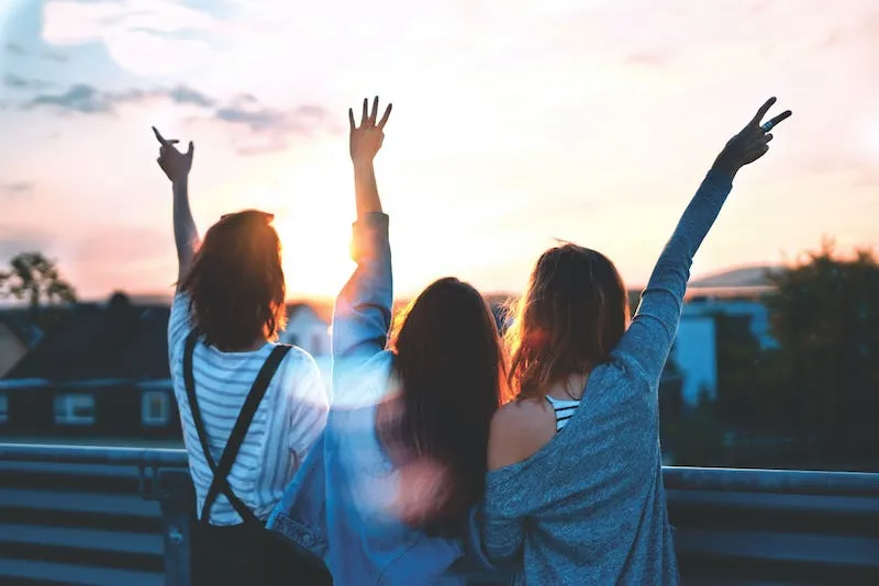 Three friends raising their arms at sunset on a rooftop, seen from behind