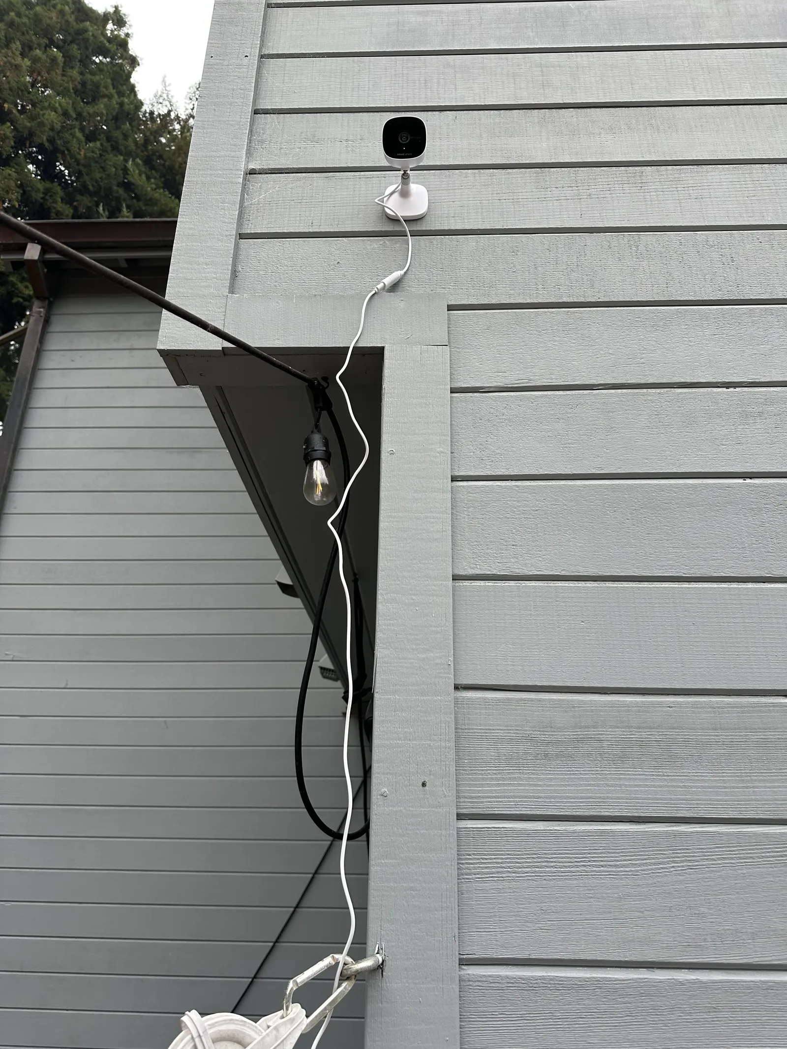 AlfredCam Plus mounted high on grey siding with its power cable running down the wall.