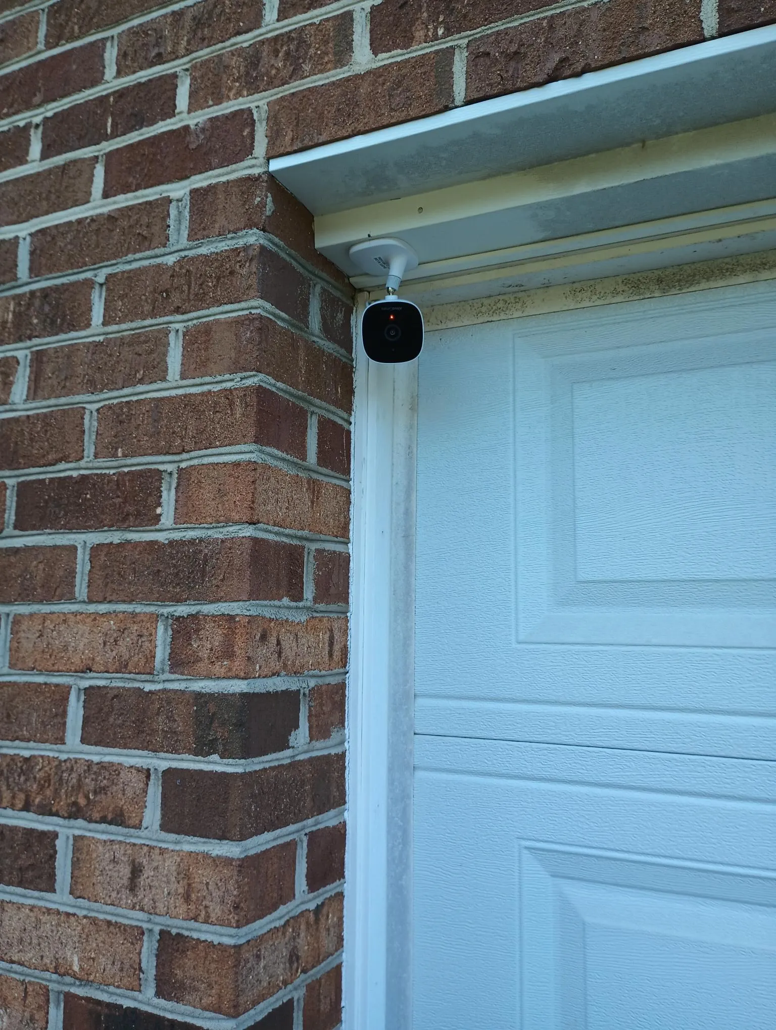 AlfredCam Plus mounted under the eave of a brick wall next to a garage door.