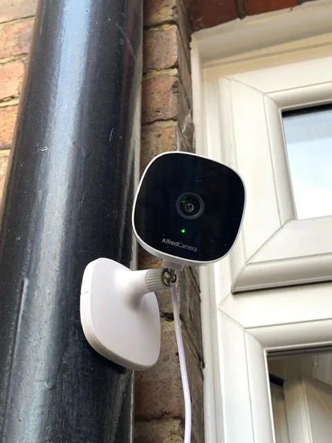 AlfredCam Plus mounted on a black water pipe outside a house.